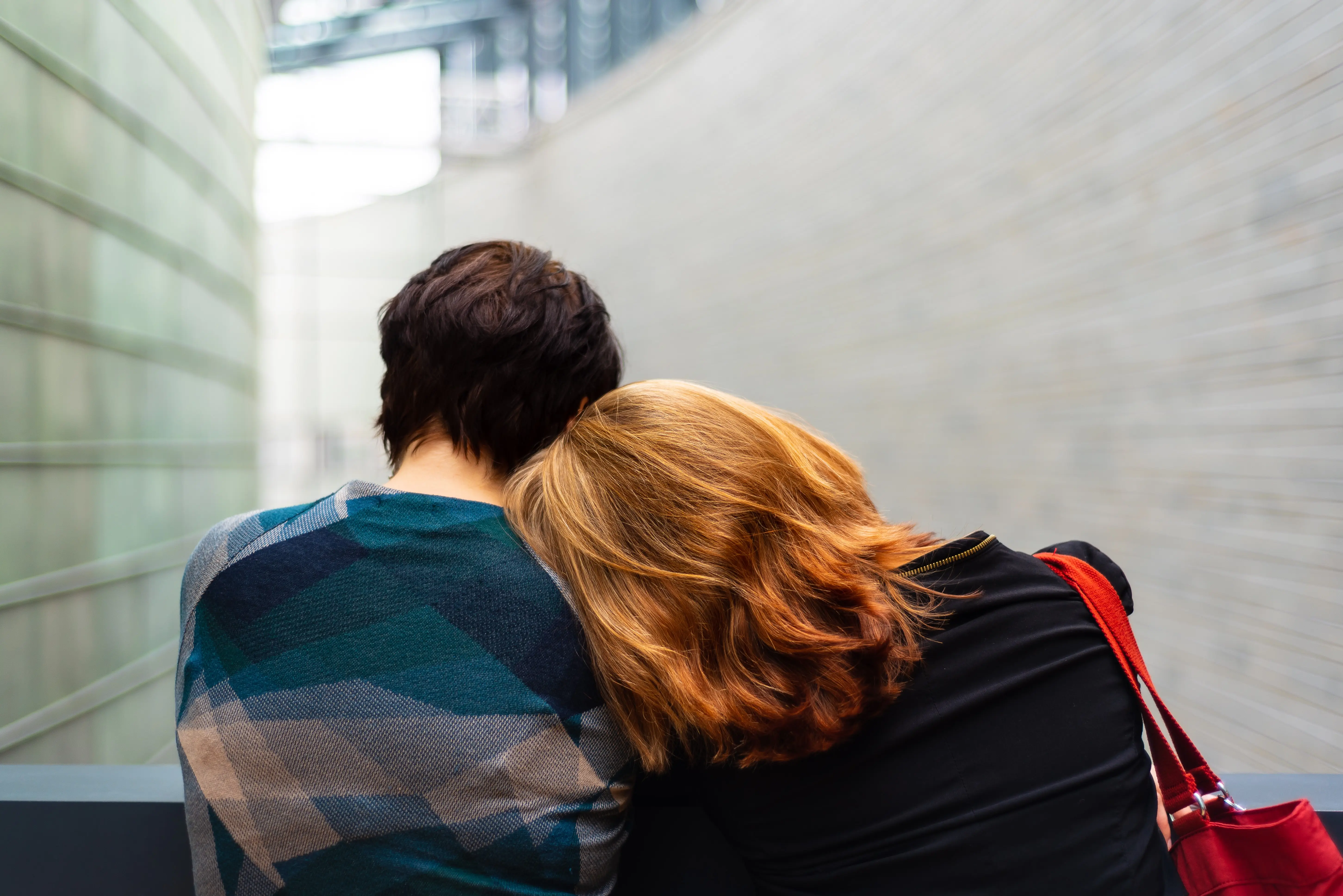 The backs of two people sitting down next to each other, the person on the right is leaning their head on the other's shoulder